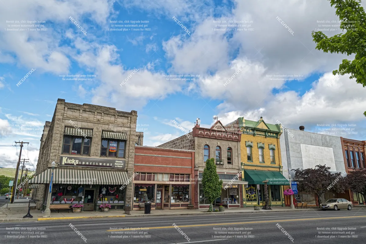 A vibrant street scene featuring colorful building facades, a local sportswear store, street trees, and an inviting sidewalk café area. The image captures the lively, walkable atmosphere of Baker City's downtown.