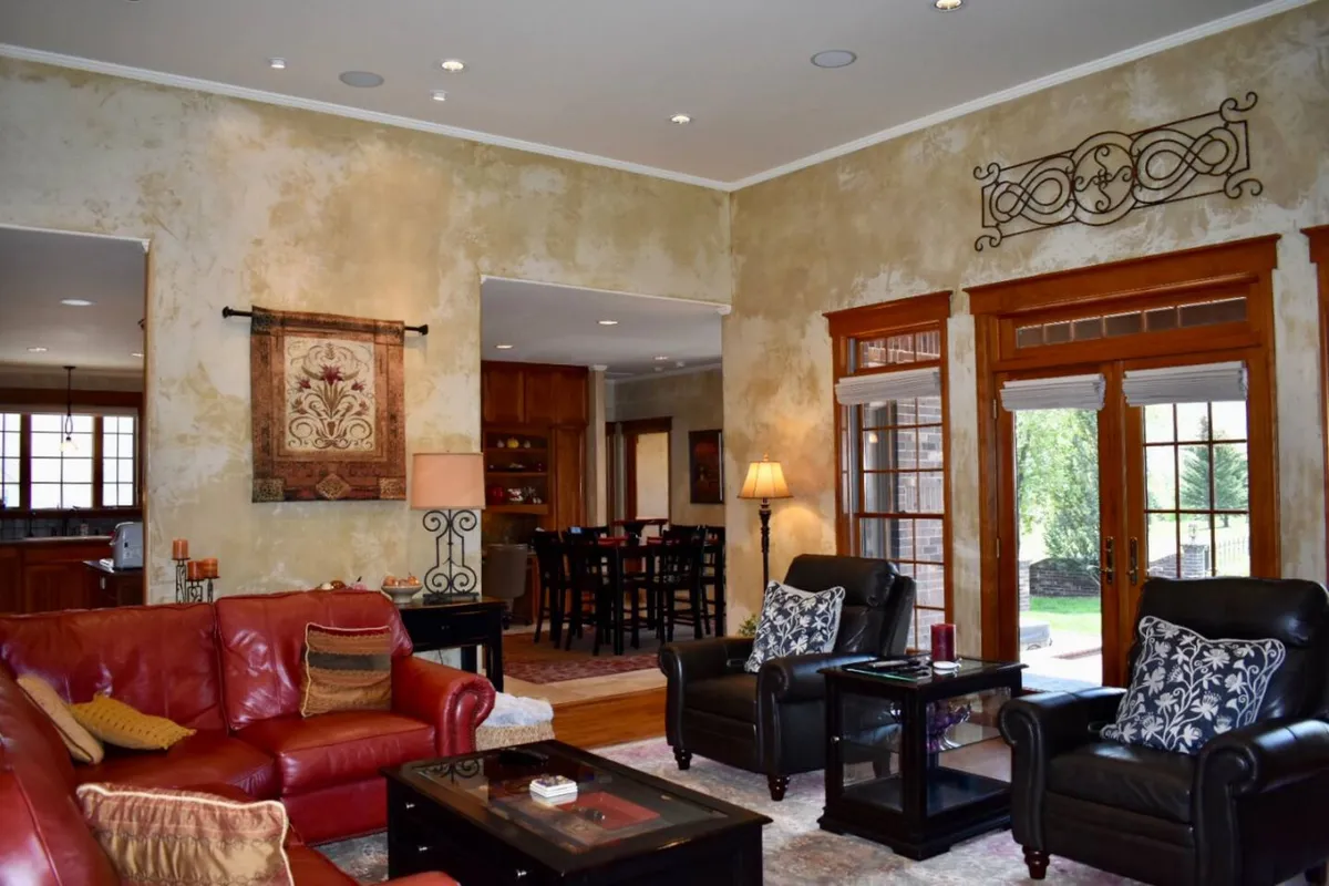 An expansive interior view featuring a red leather sofa, black leather chairs, and a dining area visible through an archway. Large windows fill the space with natural light, creating a cozy yet sophisticated setting for dining and entertaining.