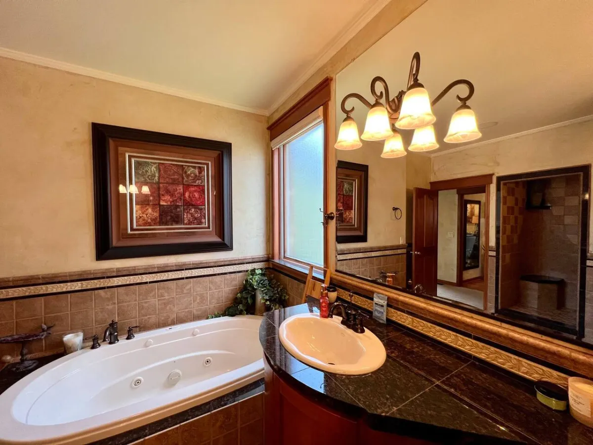 A serene bathroom scene with a jetted Jacuzzi tub, elegant sink, framed artwork, and a window allowing natural light to brighten the space. The tub is prominent, emphasizing spa-like indulgence.