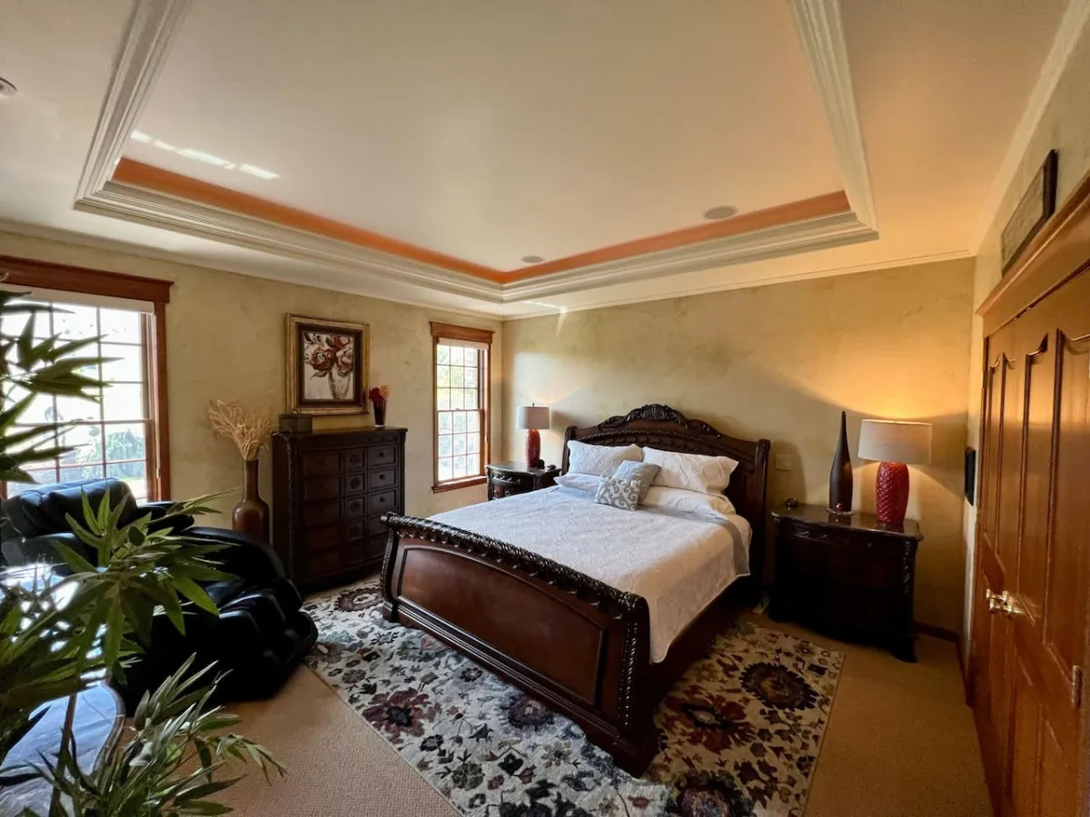 A spacious bedroom featuring a large ornate bed, cozy nightstands, a decorative rug, and windows that flood the room with natural light. The bed is outfitted with plush bedding, inviting a restful atmosphere ideal for relaxation and reading.