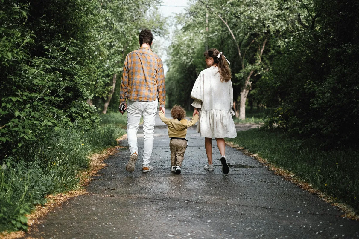 A family walks together down a tree-lined path, with a child holding the parents' hands. The scene is outdoors in a peaceful, green area, suggesting a leisurely stroll or exploration of the neighborhood. The mood is joyful and serene, focusing on connection and togetherness.