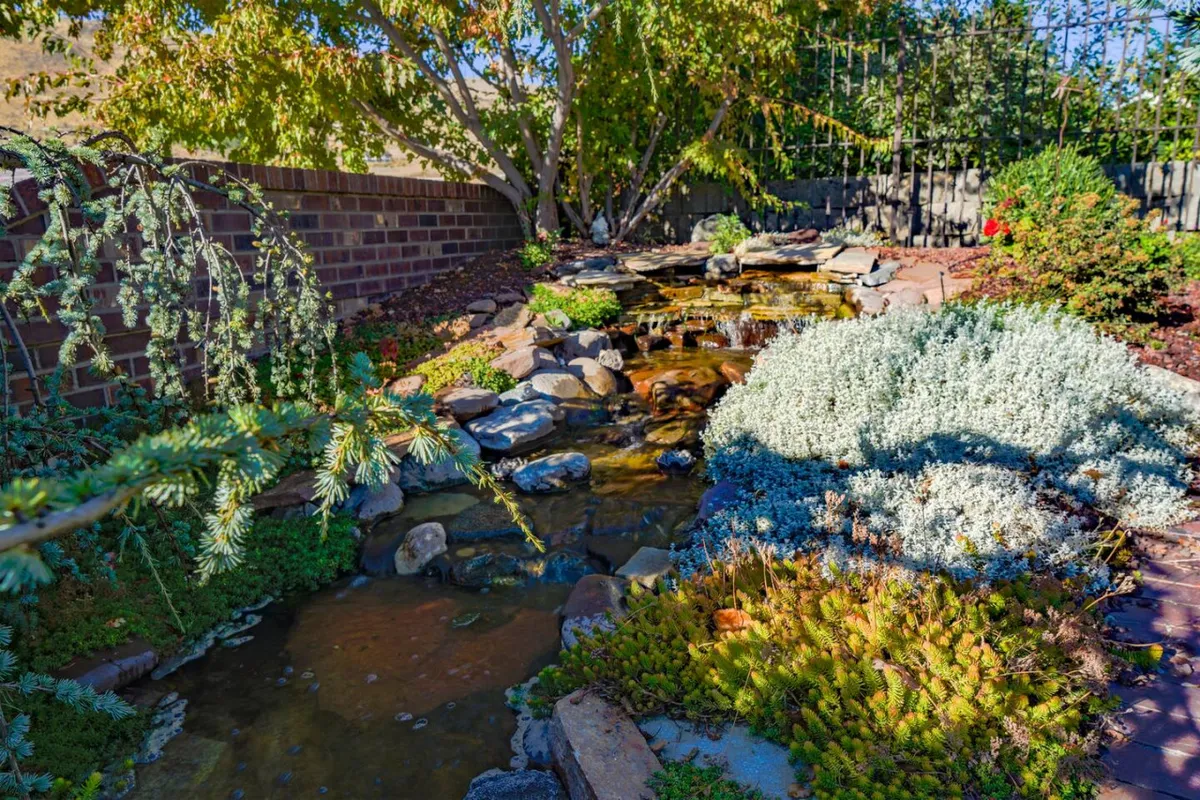 A serene water feature winds through lush gardens, bordered by a stone pathway and surrounded by various plants. The tranquil setting invites relaxation, reflection, and time spent outdoors enjoying nature’s gentle sounds.