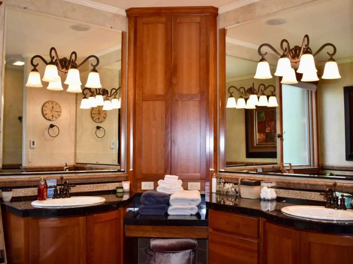 A bathroom with double vanity sinks, mirrored walls, decorative lighting fixtures, neatly stacked towels, and tasteful artwork, all bathed in gentle morning light. The space feels both elegant and functional.