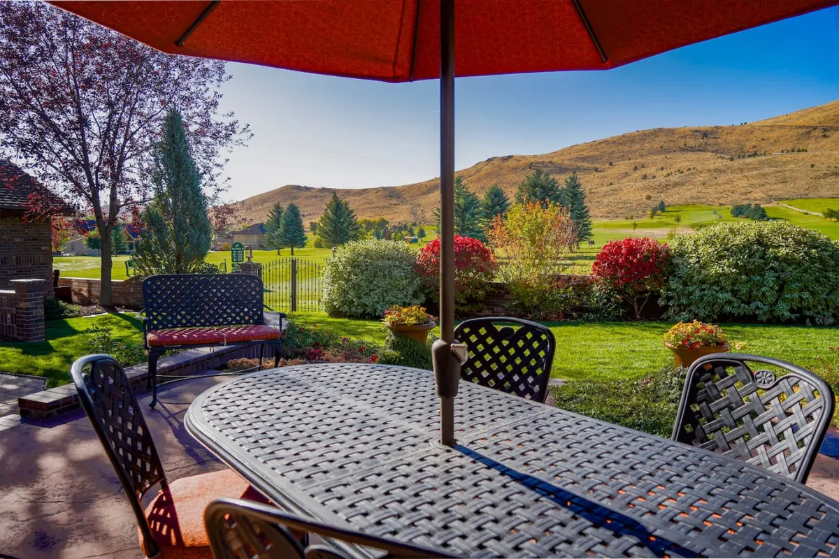 An inviting outdoor dining table shaded by an umbrella is set within a landscaped area, with a scenic landscape view in the background. The setting is ideal for al fresco dining, gathering with friends and family, and celebrating special occasions under the open sky.